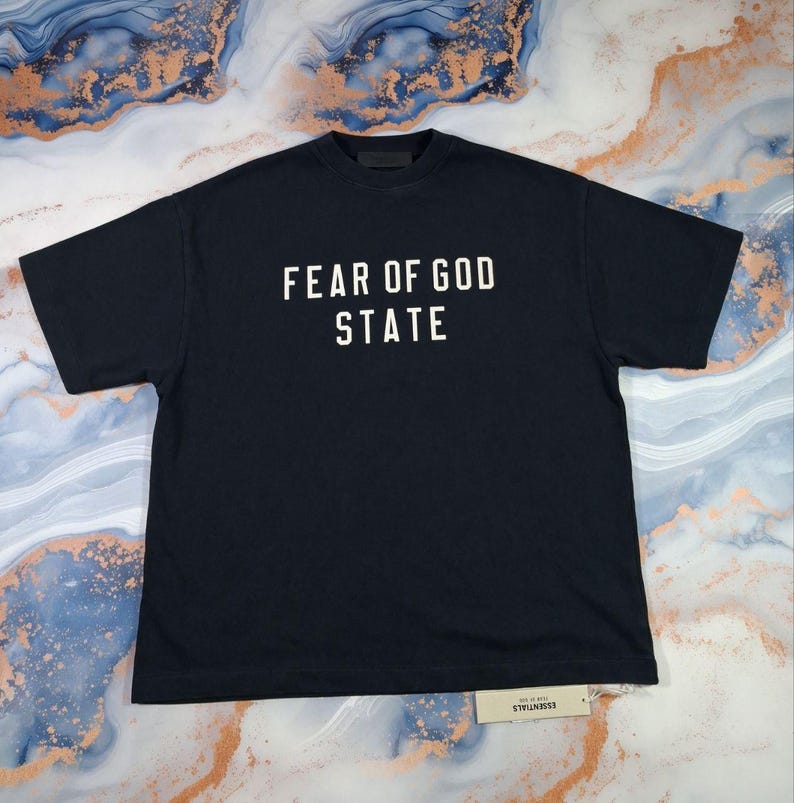 May include: A navy blue t-shirt with the words "FEAR OF GOD STATE" printed in white block letters. The shirt has a crew neck and short sleeves. A small tag is attached to the bottom of the shirt.