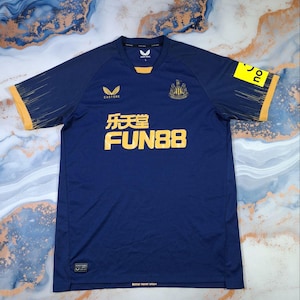 K&ouml;nnte beinhalten: Marineblaues Fu&szlig;balltrikot mit gelben Akzenten an &Auml;rmeln und Kragen. Das Trikot zeigt das Castore-Logo, ein Teamwappen und den Text "FUN88" in Gelb. Die &Auml;rmel haben ein dezentes Streifendesign. Der Text "Better never stops" steht unten.