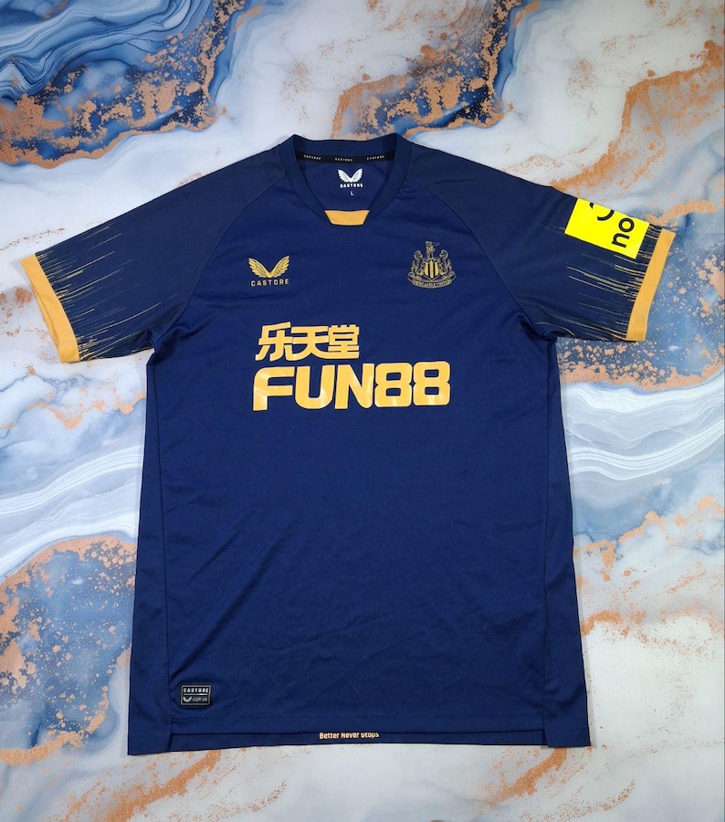 K&ouml;nnte beinhalten: Marineblaues Fu&szlig;balltrikot mit gelben Akzenten an &Auml;rmeln und Kragen. Das Trikot zeigt das Castore-Logo, ein Teamwappen und den Text "FUN88" in Gelb. Die &Auml;rmel haben ein dezentes Streifendesign. Der Text "Better never stops" steht unten.