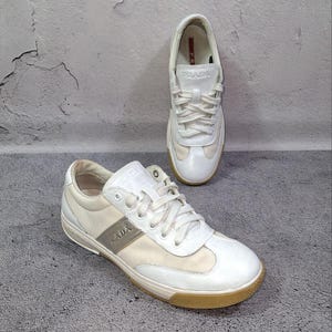 Puede incluir: Zapatillas de cuero blanco con suela marrón claro. Las zapatillas tienen cordones blancos y la marca "PRADA" es visible en el lateral. Una zapatilla está colocada delante de la otra.