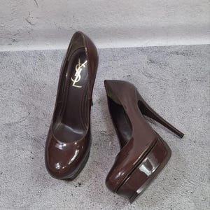 May include: A pair of brown patent leather platform pumps. The shoes have a high heel and a rounded toe. The left shoe features a gold logo. The shoes are a dark, glossy brown color.