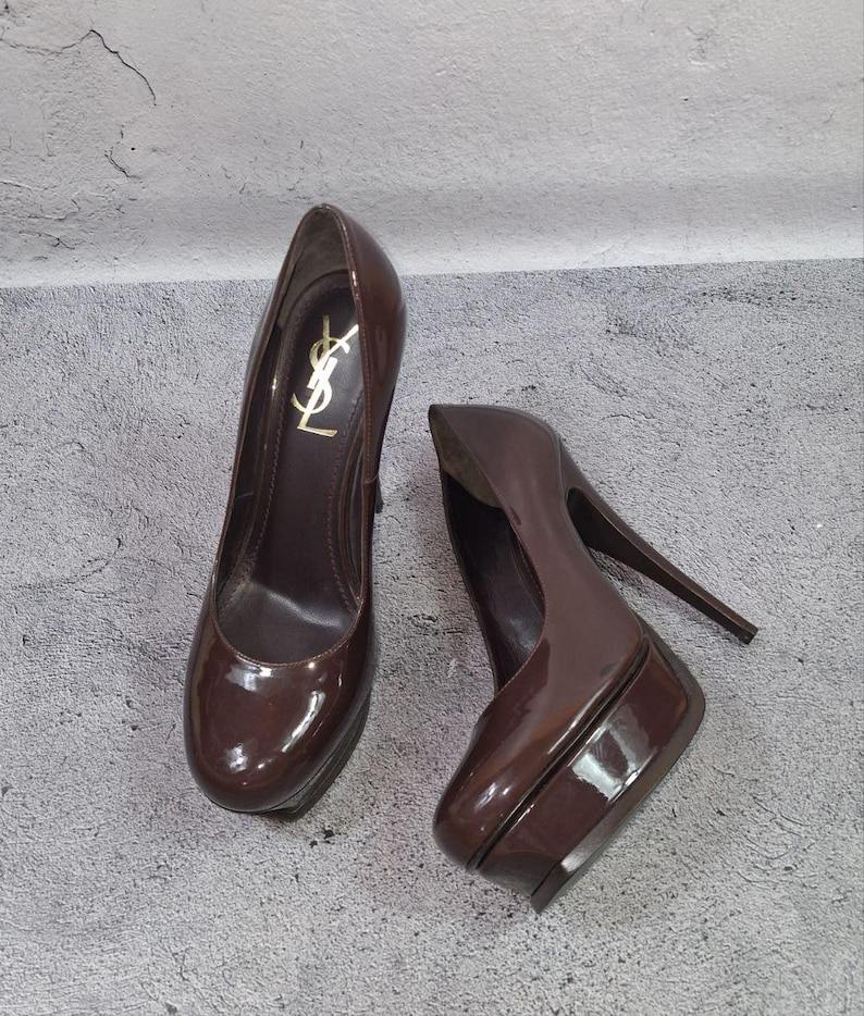 May include: A pair of brown patent leather platform pumps. The shoes have a high heel and a rounded toe. The left shoe features a gold logo. The shoes are a dark, glossy brown color.