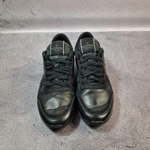 May include: Pair of black Valentino Garavani sneakers. The shoes have black laces, a black tongue, and a black sole. The Valentino logo is visible on the tongue of each shoe. The sneakers are made of leather and suede.