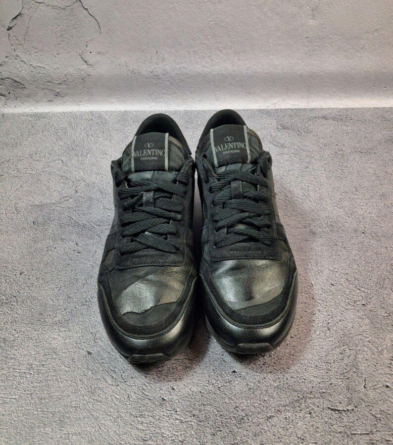 May include: Pair of black Valentino Garavani sneakers. The shoes have black laces, a black tongue, and a black sole. The Valentino logo is visible on the tongue of each shoe. The sneakers are made of leather and suede.