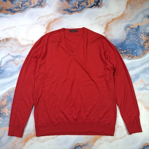 May include: A plain red V-neck jumper with long sleeves. The jumper is made of a fine knit fabric and has a ribbed hem and cuffs. The neckline is a deep V-shape.