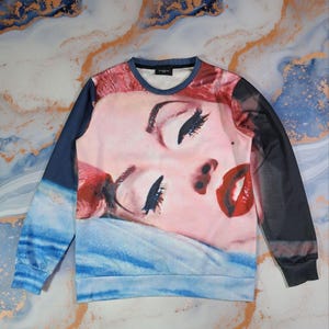 Pode incluir: Camisola de gola redonda com um close-up do rosto de uma mulher, lábios vermelhos e olhos fechados. As mangas são azul escuro e pretas. A camisola tem uma gola azul.