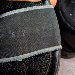 May include: Close-up of a black shoe with a mesh upper and a black strap. The strap has the numbers "5.2" and "W 7.5" written on it in white. The shoe appears to be made of leather and fabric.
