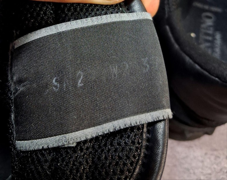 May include: Close-up of a black shoe with a mesh upper and a black strap. The strap has the numbers "5.2" and "W 7.5" written on it in white. The shoe appears to be made of leather and fabric.