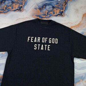May include: A navy blue t-shirt with the text "FEAR OF GOD STATE" in white block letters. The shirt is made of a soft material and has a crew neck.