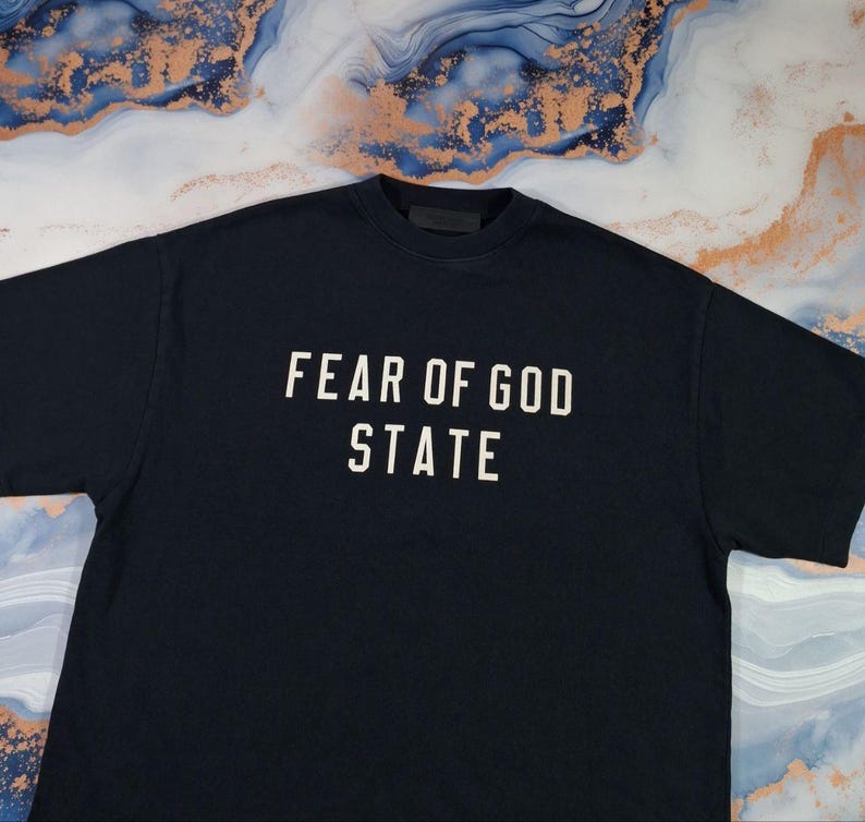 May include: A navy blue t-shirt with the text "FEAR OF GOD STATE" in white block letters. The shirt is made of a soft material and has a crew neck.