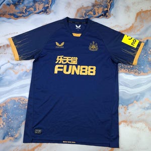 K&ouml;nnte beinhalten: Marineblaues Fu&szlig;balltrikot mit goldenen Akzenten. Das Trikot zeigt das "FUN88"-Logo in Gold, das Vereinswappen und Sponsorenlogos. Die &Auml;rmel haben goldene Verzierungen und ein dezentes Muster. Der Text "Better never stops" ist unten aufgedruckt.
