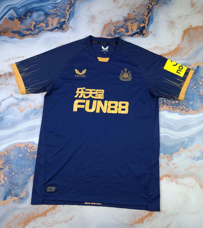 K&ouml;nnte beinhalten: Marineblaues Fu&szlig;balltrikot mit goldenen Akzenten. Das Trikot zeigt das "FUN88"-Logo in Gold, das Vereinswappen und Sponsorenlogos. Die &Auml;rmel haben goldene Verzierungen und ein dezentes Muster. Der Text "Better never stops" ist unten aufgedruckt.