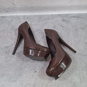 May include: A pair of brown platform high heels with stiletto heels. The shoes have a glossy finish and a rounded toe. The heels are angled and the platform is thick. The shoes are on a gray surface.