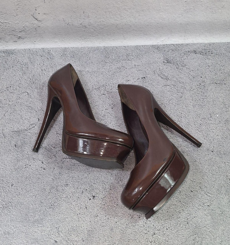 May include: A pair of brown platform high heels with stiletto heels. The shoes have a glossy finish and a rounded toe. The heels are angled and the platform is thick. The shoes are on a gray surface.