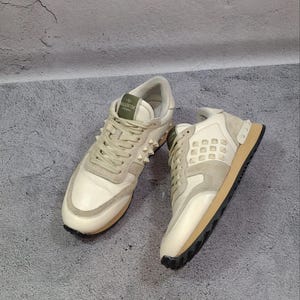 K&ouml;nnte beinhalten: Ein Paar cremewei&szlig;e und beigefarbene Sneaker mit Schn&uuml;rverschluss, einem gr&uuml;nen Zungenetikett mit dem Text Valentino Garavani und einer Seitenpartie, die mit kleinen pyramidenf&ouml;rmigen Nieten verziert ist. Die Schuhe haben eine braune Sohle.