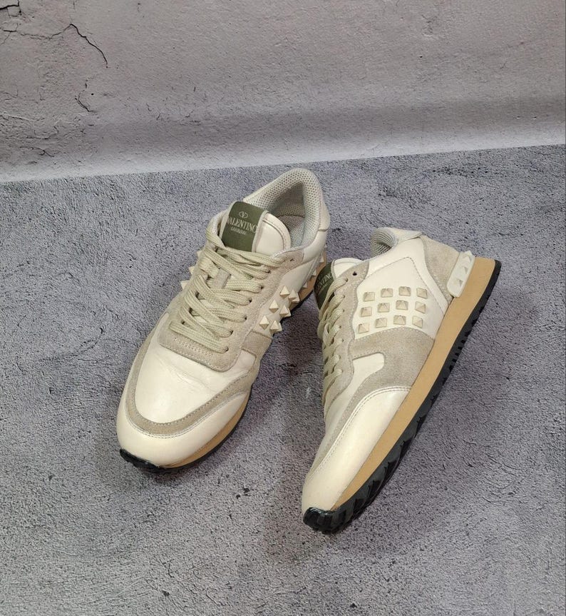 K&ouml;nnte beinhalten: Ein Paar cremewei&szlig;e und beigefarbene Sneaker mit Schn&uuml;rverschluss, einem gr&uuml;nen Zungenetikett mit dem Text Valentino Garavani und einer Seitenpartie, die mit kleinen pyramidenf&ouml;rmigen Nieten verziert ist. Die Schuhe haben eine braune Sohle.