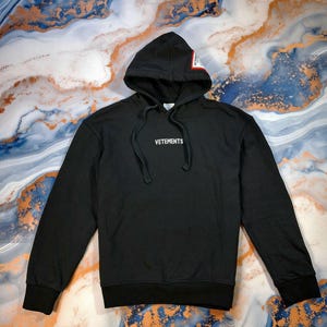 Puede incluir: Sudadera con capucha negra con la palabra "VETEMENTS" en letras blancas en el pecho. La sudadera tiene una capucha con cordón y un pequeño parche rectangular en la capucha. El fondo es un patrón veteado de azul, dorado y blanco.