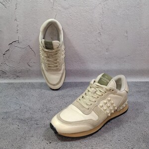K&ouml;nnte beinhalten: Ein Paar cremewei&szlig;e Sneaker mit Schn&uuml;rverschluss, Wildleder- und Ledereins&auml;tzen sowie einem Seitenteil, das mit pyramidenf&ouml;rmigen Nieten verziert ist. Die Zunge zeigt ein gr&uuml;nes Etikett mit dem Text Valentino Garavani und die Sohle ist hellbraun.