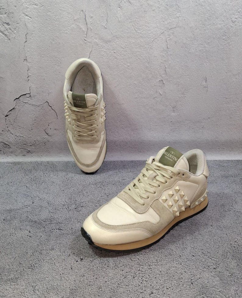 K&ouml;nnte beinhalten: Ein Paar cremewei&szlig;e Sneaker mit Schn&uuml;rverschluss, Wildleder- und Ledereins&auml;tzen sowie einem Seitenteil, das mit pyramidenf&ouml;rmigen Nieten verziert ist. Die Zunge zeigt ein gr&uuml;nes Etikett mit dem Text Valentino Garavani und die Sohle ist hellbraun.