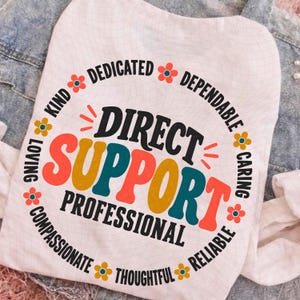 以下が含まれることがあります： DIRECT SUPPORT PROFESSIONALというテキストが大きくカラフルなレトロ調のフォントで描かれた円形のグラフィックが特徴の白いTシャツ。テキストの周囲にはKIND、DEDICATED、DEPENDABLE、CARING、RELIABLE、THOUGHTFUL、COMPASSIONATE、LOVINGという単語が配置されています。