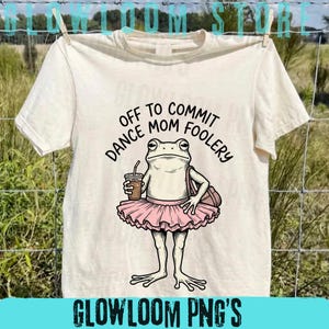 May include: Cream-colored t-shirt with a cartoon frog wearing a pink tutu, holding a coffee cup and a bag. Text on the shirt reads "DANCE MOM OFF TO COMMIT FOOLERY". The shirt is hanging on a clothesline.