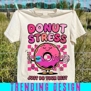 Może przedstawiać: Kremowa koszulka z grafiką różowego pączka z uśmiechniętą twarzą, trzymającego ołówek i notatnik. Tekst "DONUT STRESS" i "JUST DO YOUR BEST" są widoczne. Na dole znajduje się napis "TRENDING DESIGN".