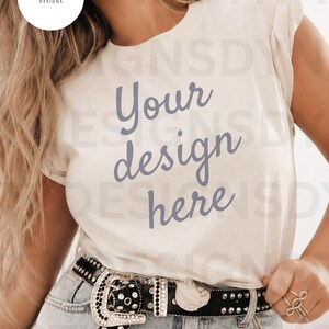 以下が含まれることがあります： ダークブルーの筆記体フォントで「Your design here」とプリントされたクリーム色のTシャツ。Tシャツはライトブルーのデニムジーンズと、シルバーのスタッズと大きな装飾バックルが付いた黒いベルトと組み合わされています。