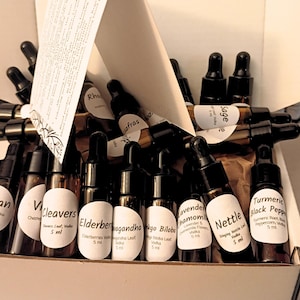 May include: A box filled with small brown glass bottles with black droppers. Each bottle has a white label with text, including "Valerian," "Cleavers," and "Nettle." The box also contains a folded paper with text.