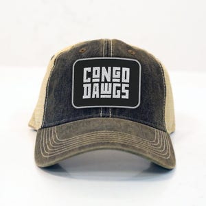 Op de afbeelding: Een baseballpet met een verweerde look in zwart en beige. De voorkant heeft een rechthoekige patch met de woorden "CONGO DAWGS" in witte blokletters. De pet heeft een gebogen klep en een mesh achterkant.