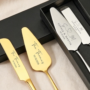 Puede incluir: Un juego de cuchillo y servidor para pastel de bodas en una caja negra. Un juego es dorado, el otro plateado. Ambos juegos están grabados con nombres y una fecha. Rosas rosas y paniculata en el fondo.
