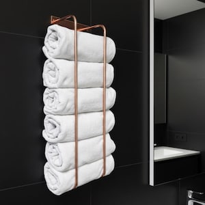 Könnte beinhalten: Ein kupferfarbener Handtuchhalter mit sechs gerollten weißen Handtüchern. Der Halter ist an einer schwarzen gefliesten Wand montiert. Ein Spiegel und ein Waschbecken sind im Hintergrund zu sehen, was auf ein Badezimmer hindeutet.