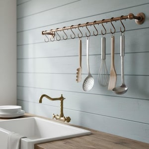 May include: A kitchen setting with a copper-toned utensil rack holding several cooking tools. These include a slotted spoon, whisk, spatula, and ladle. The backdrop is a light blue wall with horizontal wooden planks.