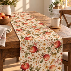 May include: A quilted table runner featuring a repeating pattern of red apples, green leaves, and small white and yellow flowers on a cream-colored background. The fabric has a diamond-stitched texture and is laid flat across a wooden table.