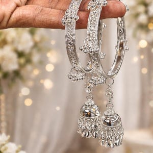 May include: Ornate silver bangles with dangling bell charms. The bangles are held up, displaying their detailed design and reflective surface. The jewellery is set against a soft, blurred background.