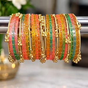 Puede incluir: Un conjunto de pulseras coloridas en tonos rosa, verde, naranja y amarillo. Cada pulsera está adornada con pequeños dijes dorados. Las pulseras se exhiben en una barra plateada, con un fondo floral borroso.