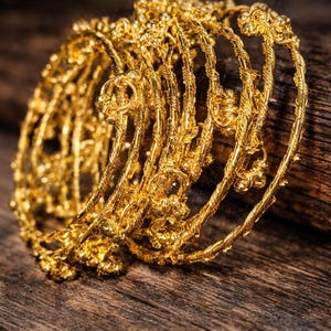 Op de afbeelding: Een verzameling gouden armbanden, ingewikkeld ontworpen met bloemen- en gedraaide touwpatronen. De armbanden zijn op elkaar gestapeld, reflecteren licht en liggen op een rustiek houten oppervlak. De sieraden hebben een warme, gouden kleur.