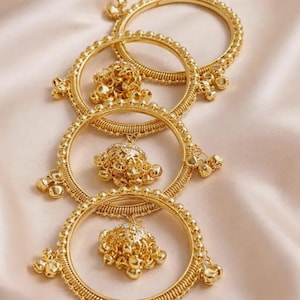Peut inclure: Quatre bracelets dorés avec des motifs perlés complexes et des breloques pendantes. Chaque bracelet présente un pendentif central orné avec des accents scintillants. Les bijoux sont présentés sur un tissu doux de couleur pêche, créant une sensation de luxe.