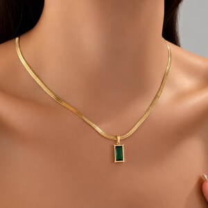 Op de afbeelding: Een goudkleurige visgraatketting met een rechthoekige hanger met een diepgroene, smaragdgeslepen edelsteen in een goudkleurig metalen frame. De ketting rust tegen de hals en toont de platte, geweven textuur van de schakels van de ketting.