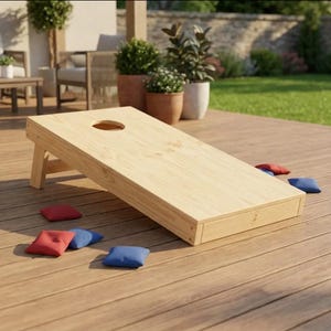 Peut inclure: Un plateau de jeu cornhole en bois clair avec un trou circulaire sur le dessus, incliné sur une terrasse en bois. Plusieurs petits sacs de haricots carrés de couleurs rouge et bleue unies sont éparpillés sur la surface de la terrasse.