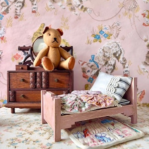 May include: A miniature wooden doll bed painted pink, featuring a white pillow with lace trim, a patchwork quilt, and a small teddy bear sitting on a dark wood dresser with a mirror, all set against a pink wallpaper with a kitten pattern.