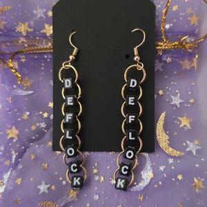 May include: A pair of dangle earrings featuring gold-tone metal jump rings linked together in a vertical chain. Each earring incorporates black square beads with white lettering that spells out the word DEFLOCK in a vertical orientation.
