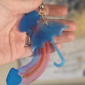 May include: A keychain featuring a blue umbrella and a rainbow design. The umbrella has a curved handle and is connected to a rainbow-shaped piece with pink, orange, and purple hues. The keychain is attached to a gold-colored clasp.