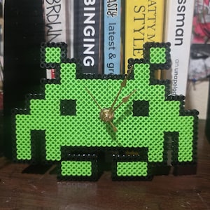 Puede incluir: Un reloj alienígena pixelado verde y negro hecho de cuentas. El reloj tiene manecillas doradas y recuerda a un personaje clásico de videojuego. El reloj se exhibe sobre una superficie de madera.