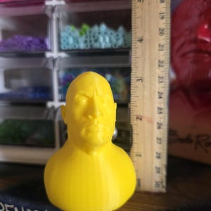 Może przedstawiać: Żółty gipsowy odlew głowy mężczyzny, o wysokości około 7.6 cm, stoi obok linijki. Gipsowy odlew stoi na ciemnej powierzchni z napisem "PENANCE" i "penance". Kolorowe koraliki widoczne w tle.