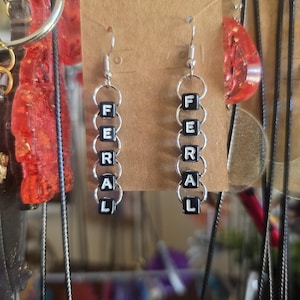Op de afbeelding: Een paar bungelende oorbellen met het woord "FERAL" gespeld in zwarte letterkralen. De oorbellen hangen aan zilverkleurige ringen en haken. Ze worden gepresenteerd op een bruine kaart.