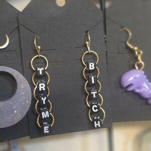 Può includere: Orecchini a cerchio color oro con perline nere con le scritte "TRY ME" e "BITCH". Gli orecchini pendono da un'etichetta nera. Un ciondolo a forma di dinosauro viola è visibile a destra.