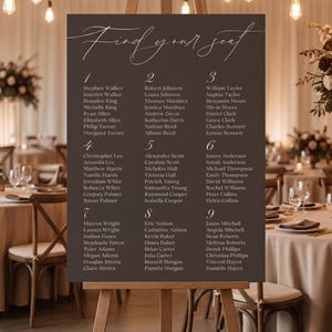 May include: A dark brown seating plan on an easel, with the words "Find your seat" in elegant calligraphy. Guest names are listed in numbered columns. The background shows a blurred image of a wedding reception, with tables and decorations.