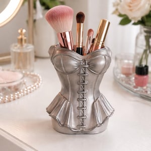 Può includere: Un portapennelli per trucco a forma di corsetto color argento pieno di vari pennelli per il trucco. Il supporto ha un design dettagliato con un davanti allacciato e un fondo a balze. I pennelli includono un grande pennello rosa e diversi pennelli più piccoli con manici dorati e neri.
