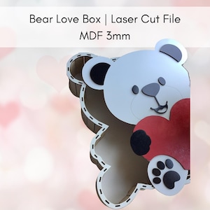 Könnte beinhalten: Eine lasergeschnittene Holzbox in Bärenform, die ein rotes Herz hält. Der Bär hat ein weißes Gesicht, eine graue Nase und einen schwarzen Pfotenabdruck. Der Text oben lautet "Bear Love Box | Laser Cut File MDF 3mm."