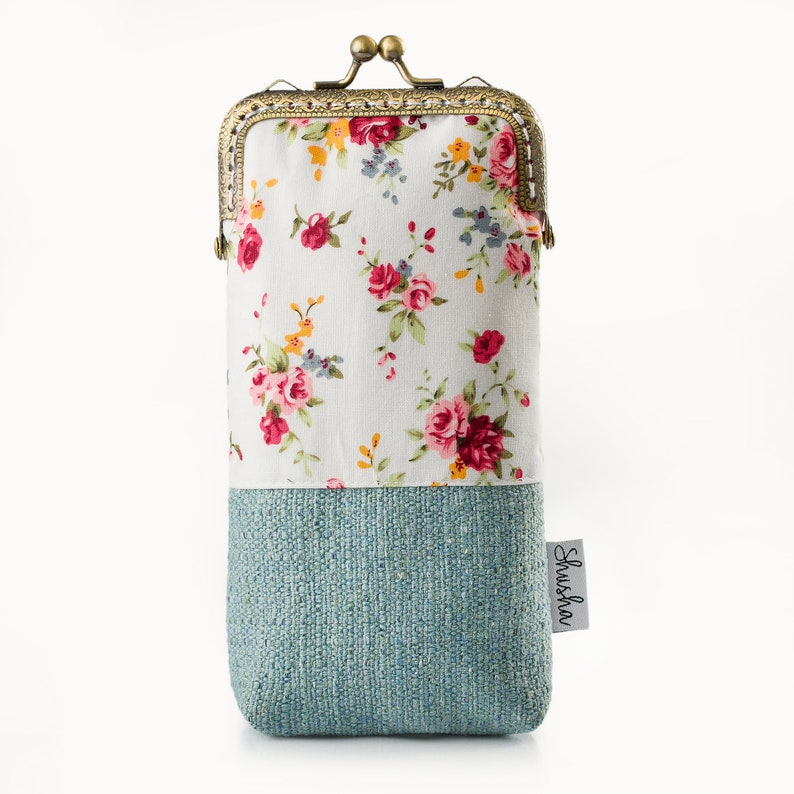 May include: A rectangular eyeglasses case with a floral pattern on the top half and a textured teal fabric on the bottom. The case has a vintage-style clasp and a small tag that reads "Shusha".